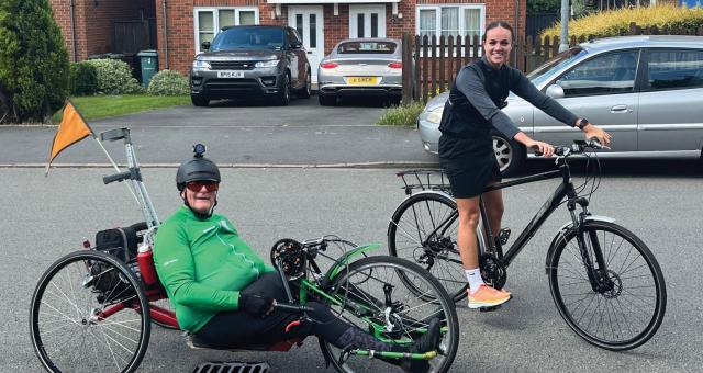 A man on a handcycle with a woman on a hybrid bike. He's wearing a green cycling jersey and black leggings. She's wearing black cycling gear and trainers.