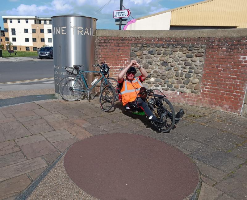 A man on a handcycle has his hands in the air in an expression of celebration. He's wearing black leggings and an orange hi-vis vest. Behind him a touring bike is leaning against a metal post that reads Transpennine Trail