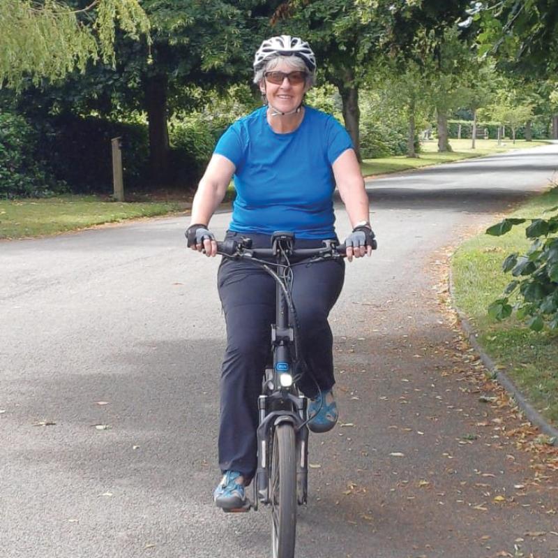 A woman is riding an e-bike towards the camera. She's wearing a blue T-shirt and trousers. She's smiling at the camera
