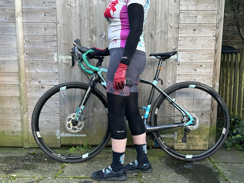 A woman in cycling jersey and shorts with Rapha arm and knee warmers is standing in front of a Felt road bike leaning against a garden shed