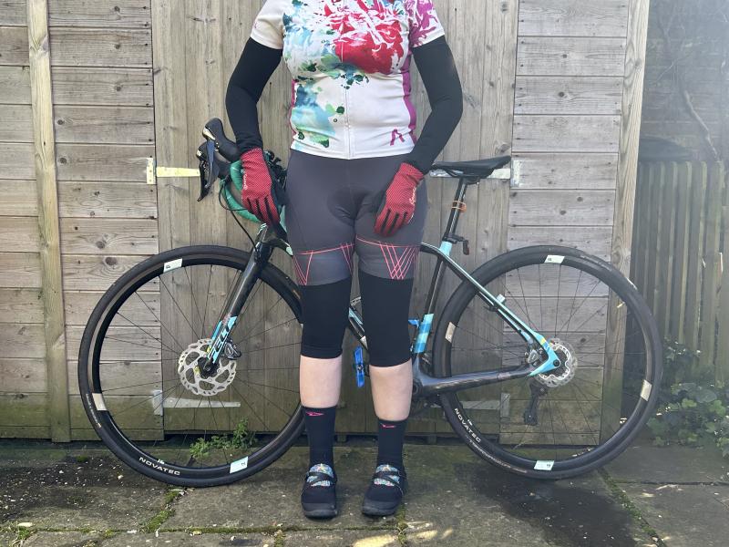 A woman in cycling jersey and shorts with Sportful arm and knee warmers is standing in front of a Felt road bike leaning against a garden shed