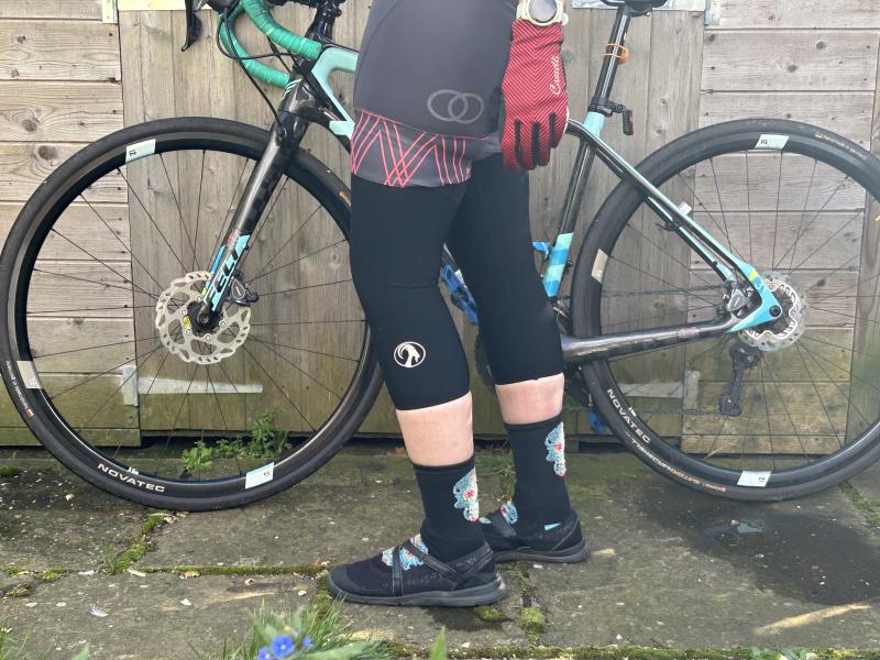 A woman in cycling jersey and shorts with Stolen Goat knee warmers is standing in front of a Felt road bike leaning against a garden shed